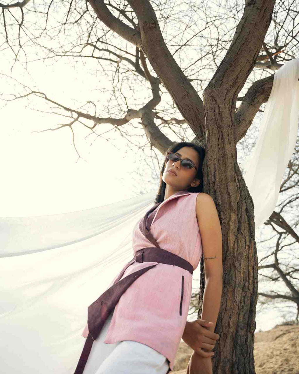 A woman wearing a pink sleeveless wrap-style top with a dark purple belt and white pants leans against a tree outdoors, with white fabric draped in the background. eco print, natural fabric, kala cotton, mauve, reversible.